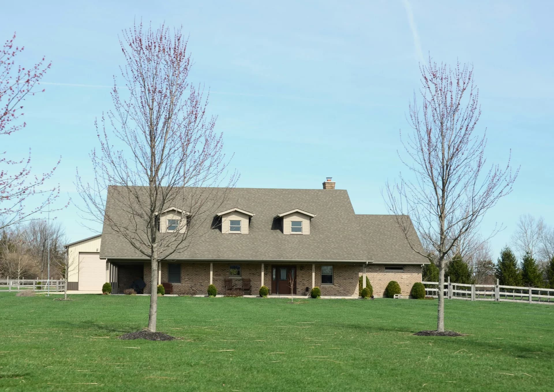 Large brick house with dormers on green lawn, framed by bare trees with white fence in rural setting