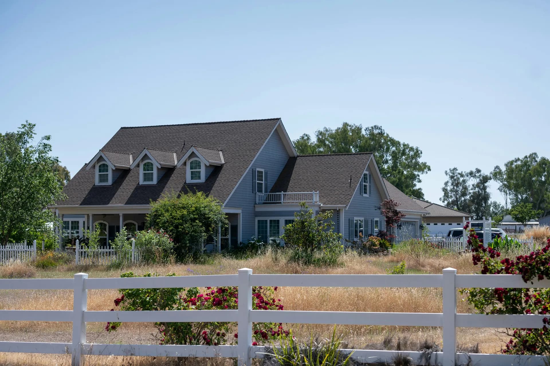Blue Cape Cod house with dormer windows behind white picket fence, surrounded by gardens and golden fields