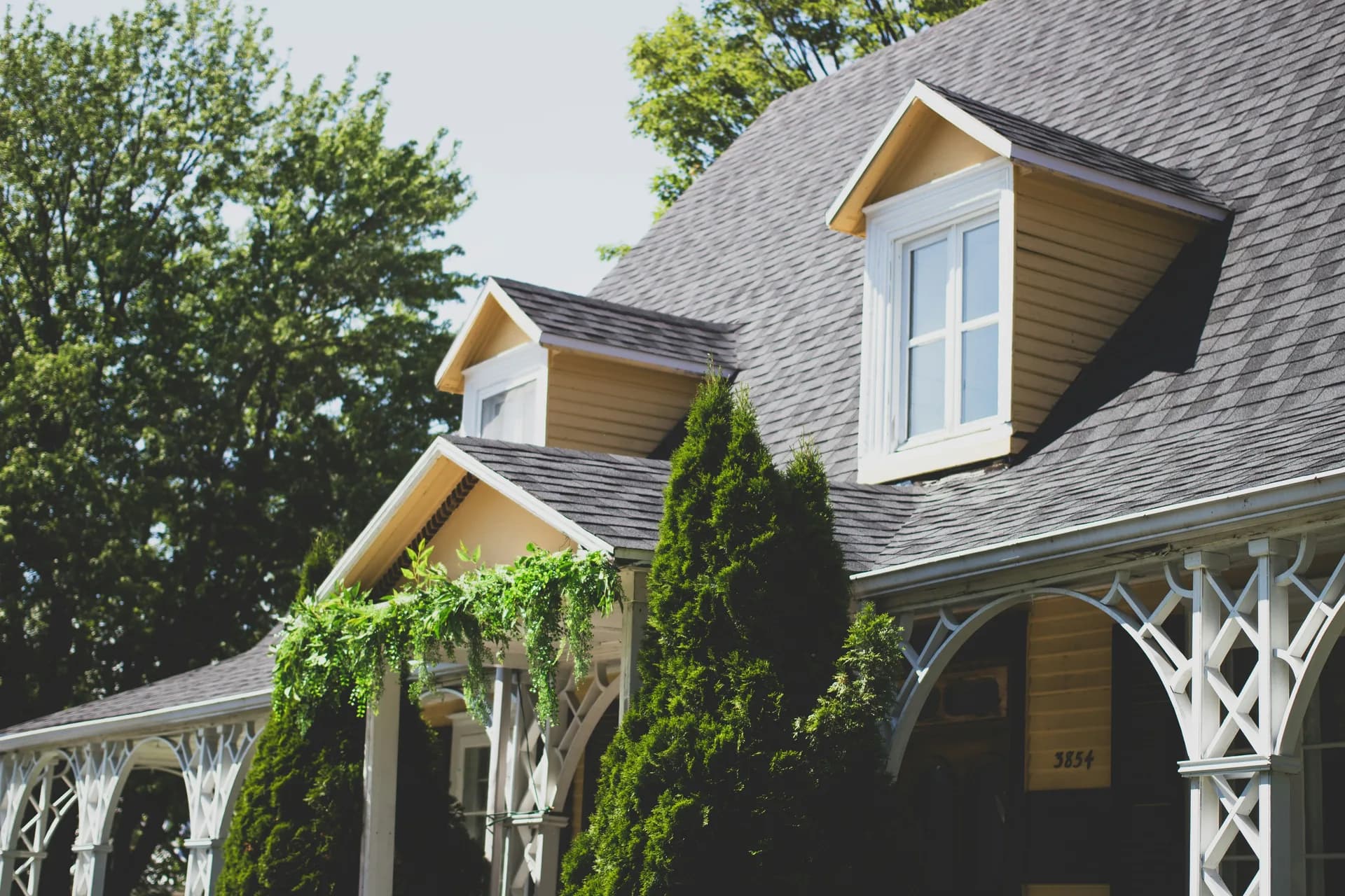 Elegant two-story house with gray roof, wood siding, white decorative porch, and dormer windows surrounded by trees