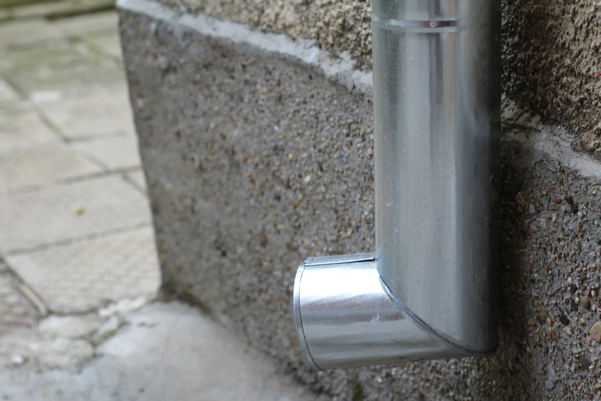Metal downspout with curved elbow joint attached to concrete wall near paved walkway