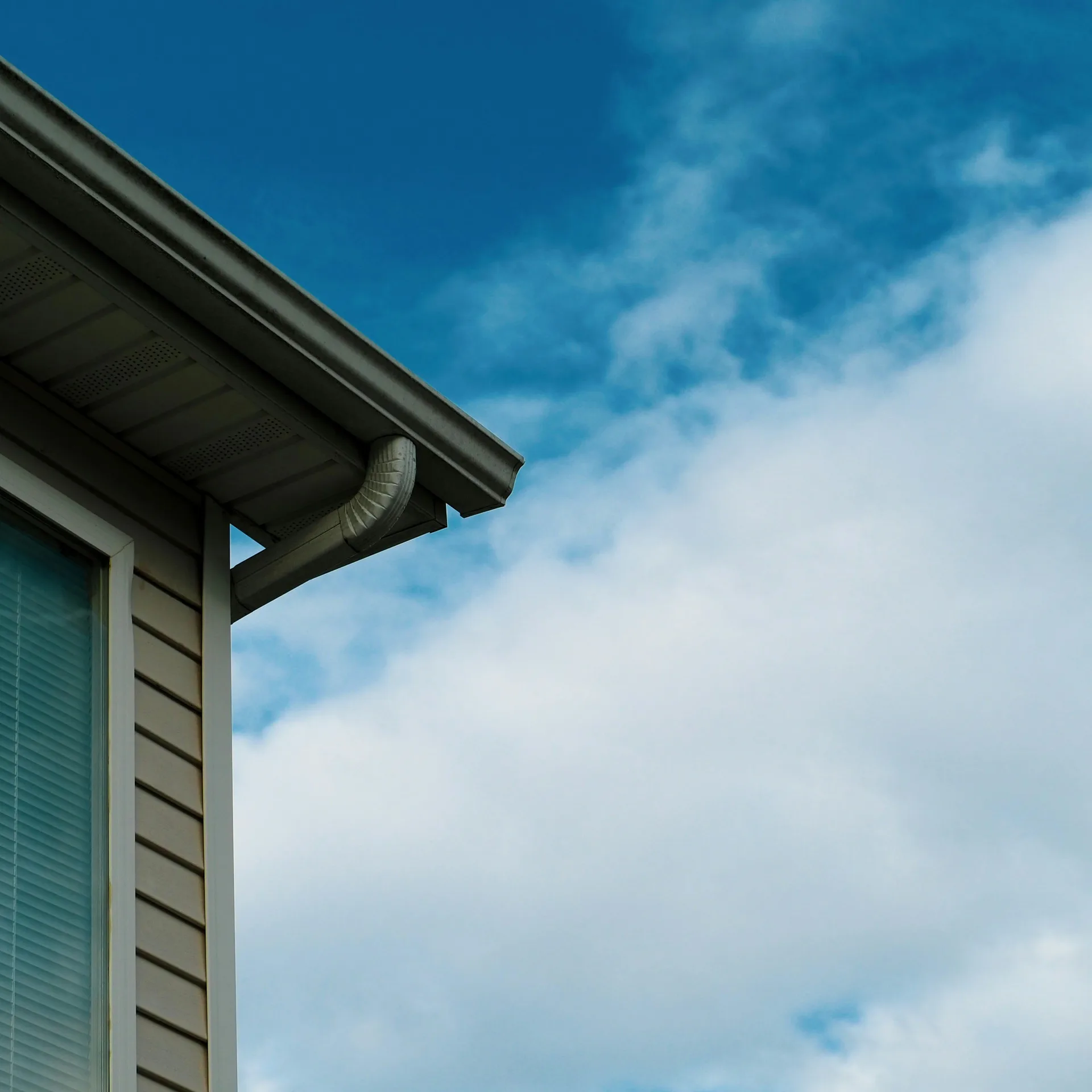 Corner of house with beige siding and dark gutters against blue cloudy sky