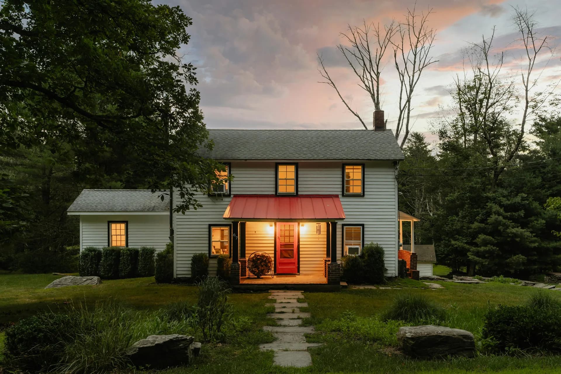 White farmhouse with red metal roof and glowing windows at sunset, surrounded by trees and green lawn