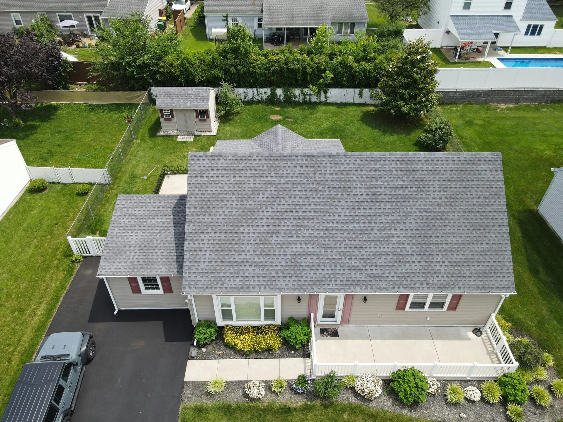 Aerial view of suburban ranch home with gray roof, beige siding, red shutters, and landscaped yard