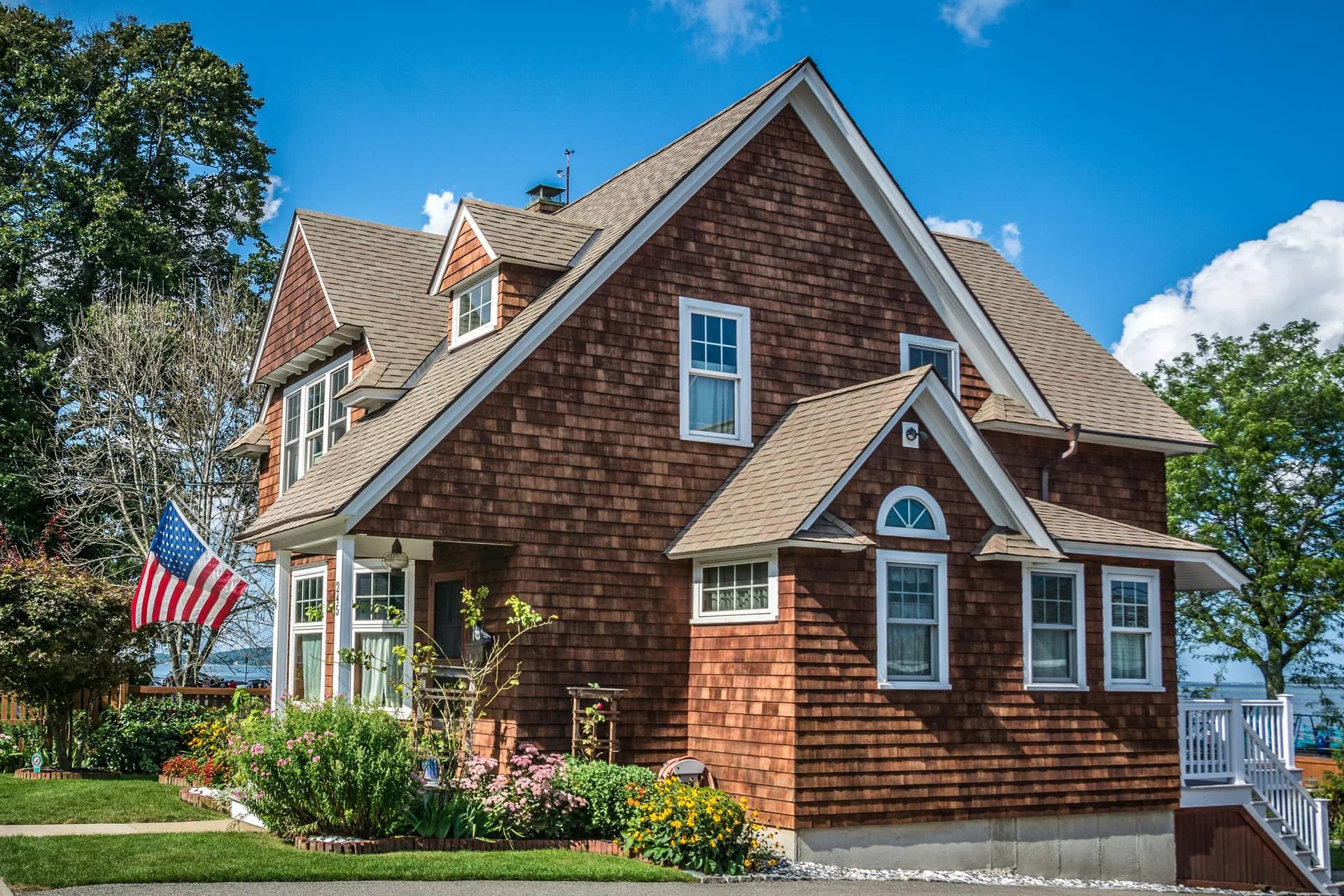 Brown cedar shake house with white trim, American flag, flower garden, and blue sky