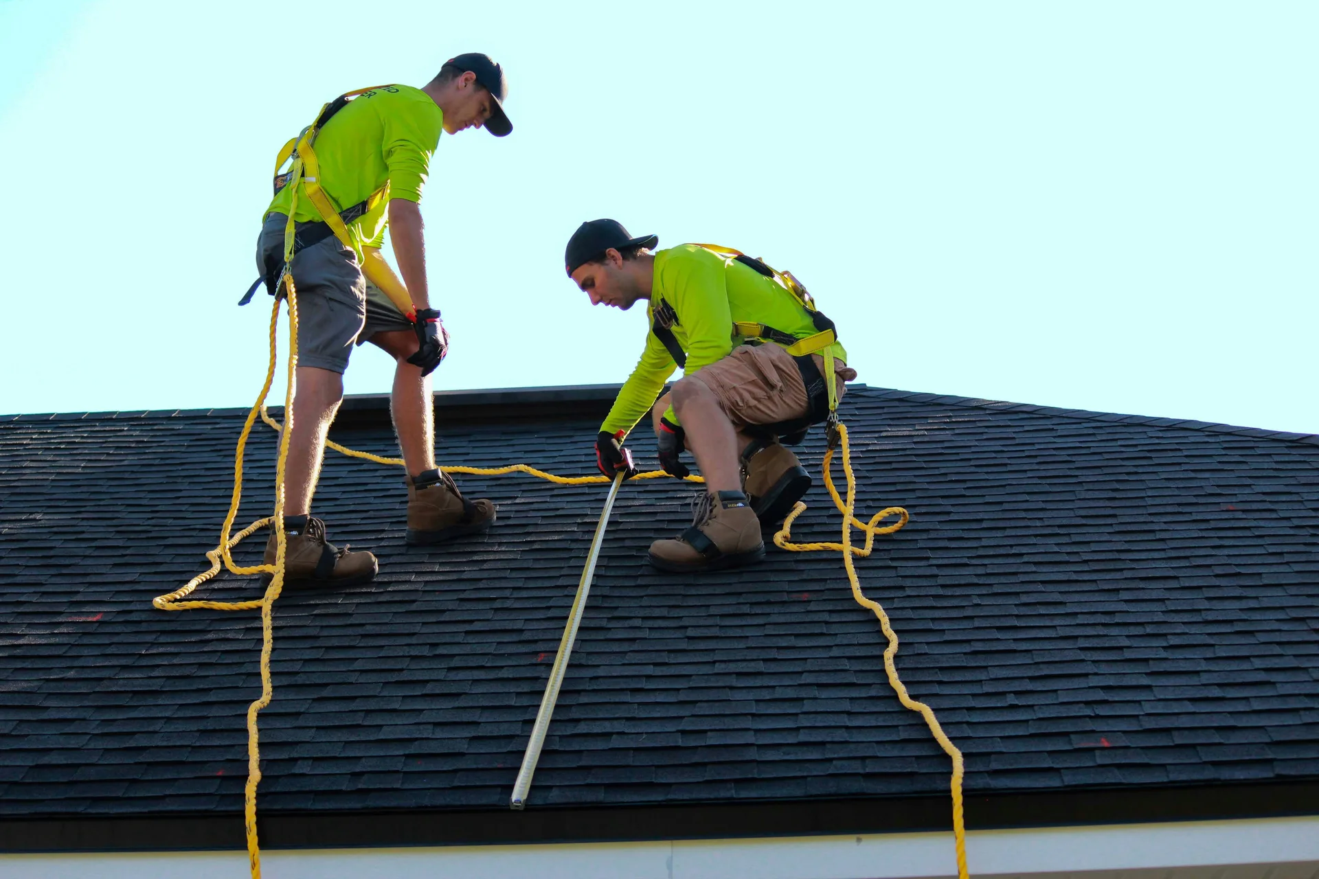 Two roofers in safety gear working on a shingled roof with yellow safety ropes