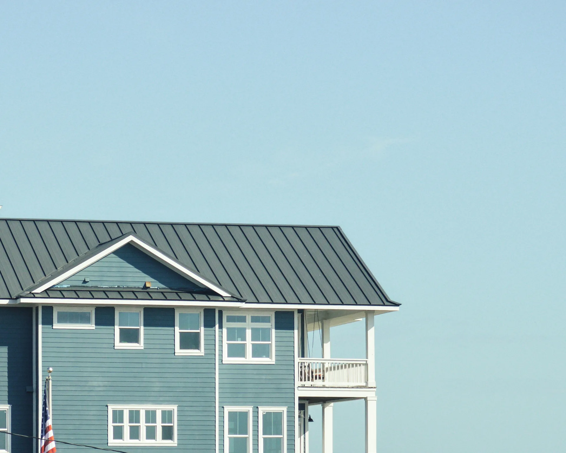 Green wooden house with white trim and metal roof against pale blue sky, featuring upper balcony and American flag