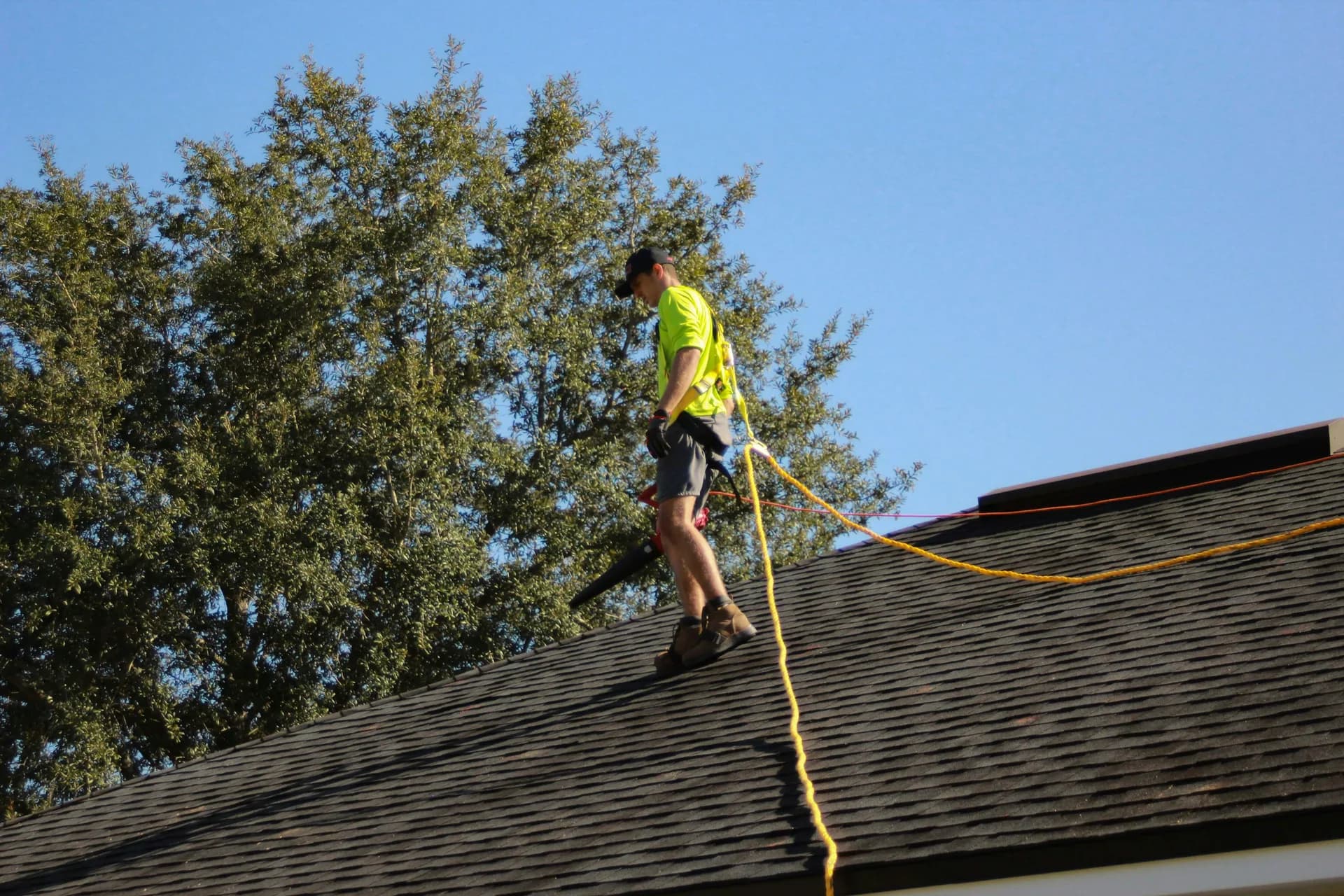 Roofing worker in yellow safety shirt standing on shingled roof with safety rope on clear day