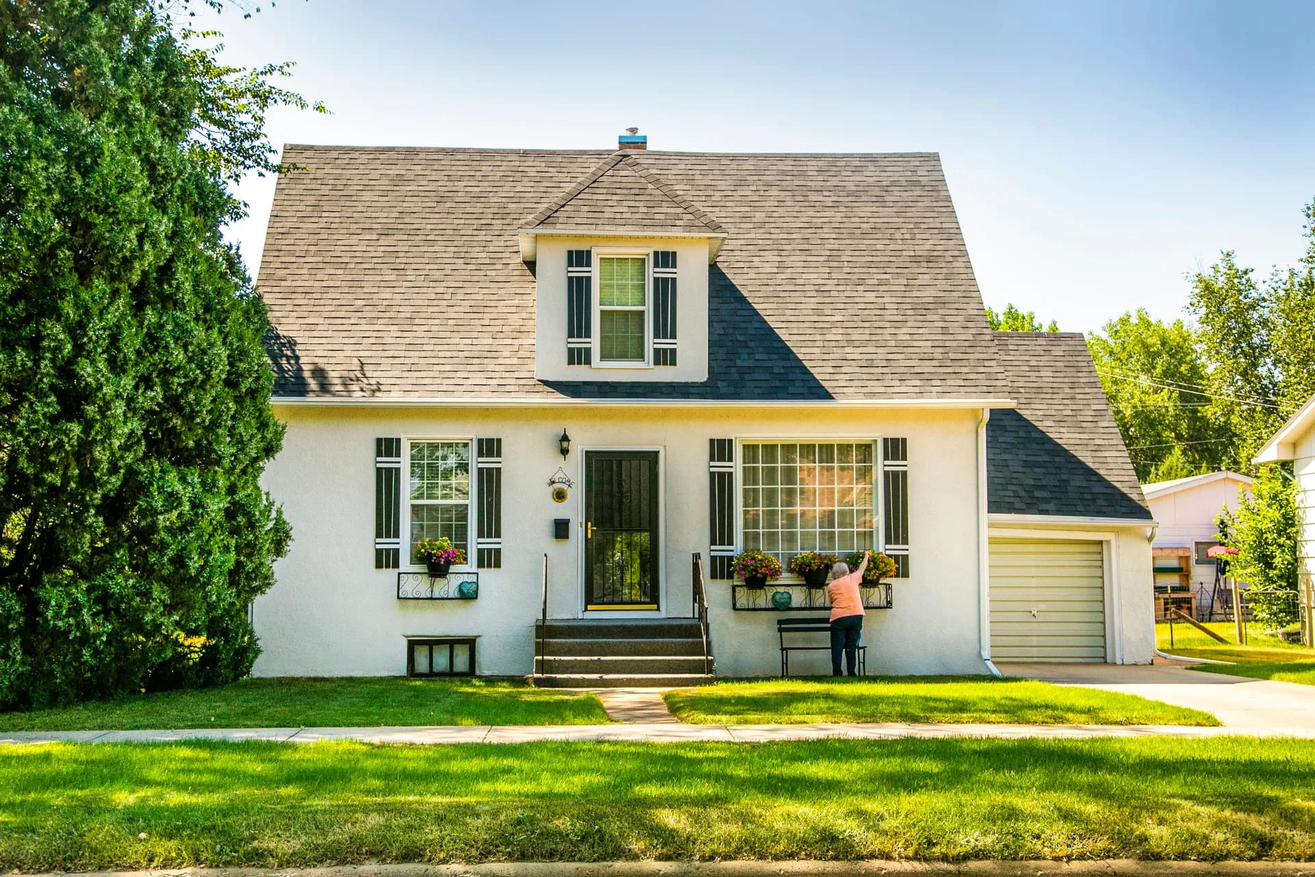 White Cape Cod style house with dormer window, dark shutters, flower boxes, and green lawn on sunny day