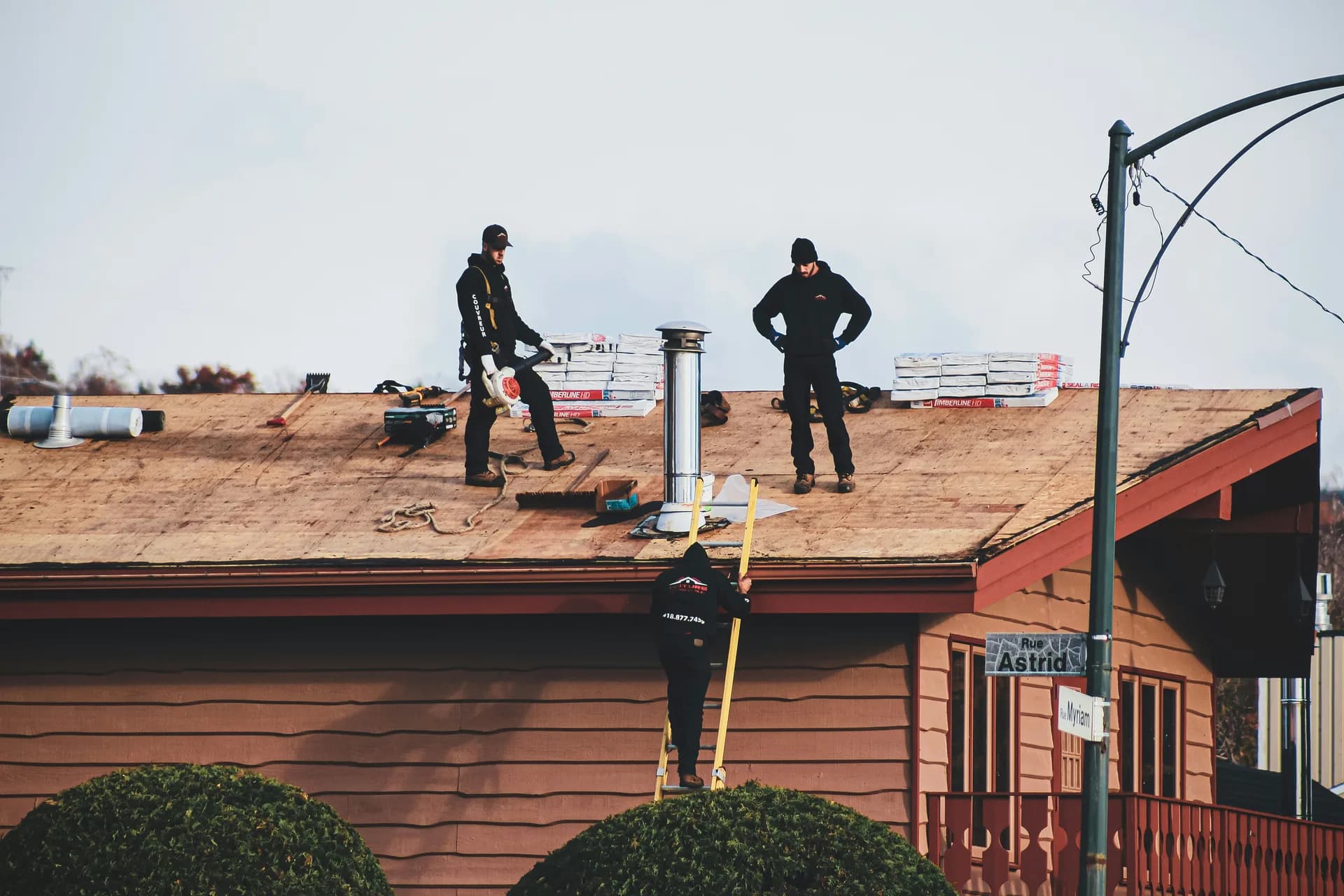 Three workers installing or repairing a roof on a brown wooden building, one on ladder, two on rooftop