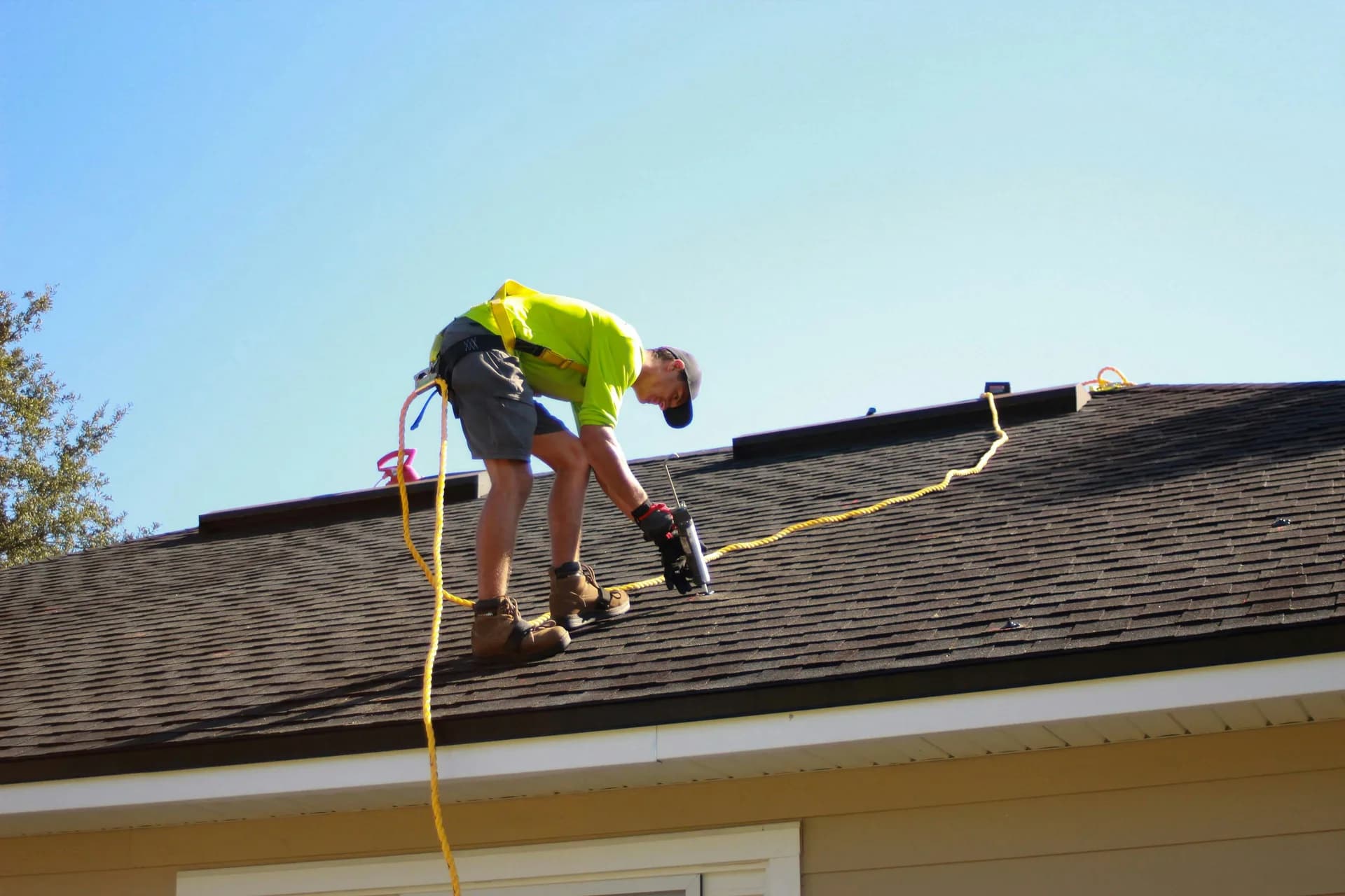 Roofer in yellow safety shirt working on asphalt shingle roof with safety harness and rope equipment