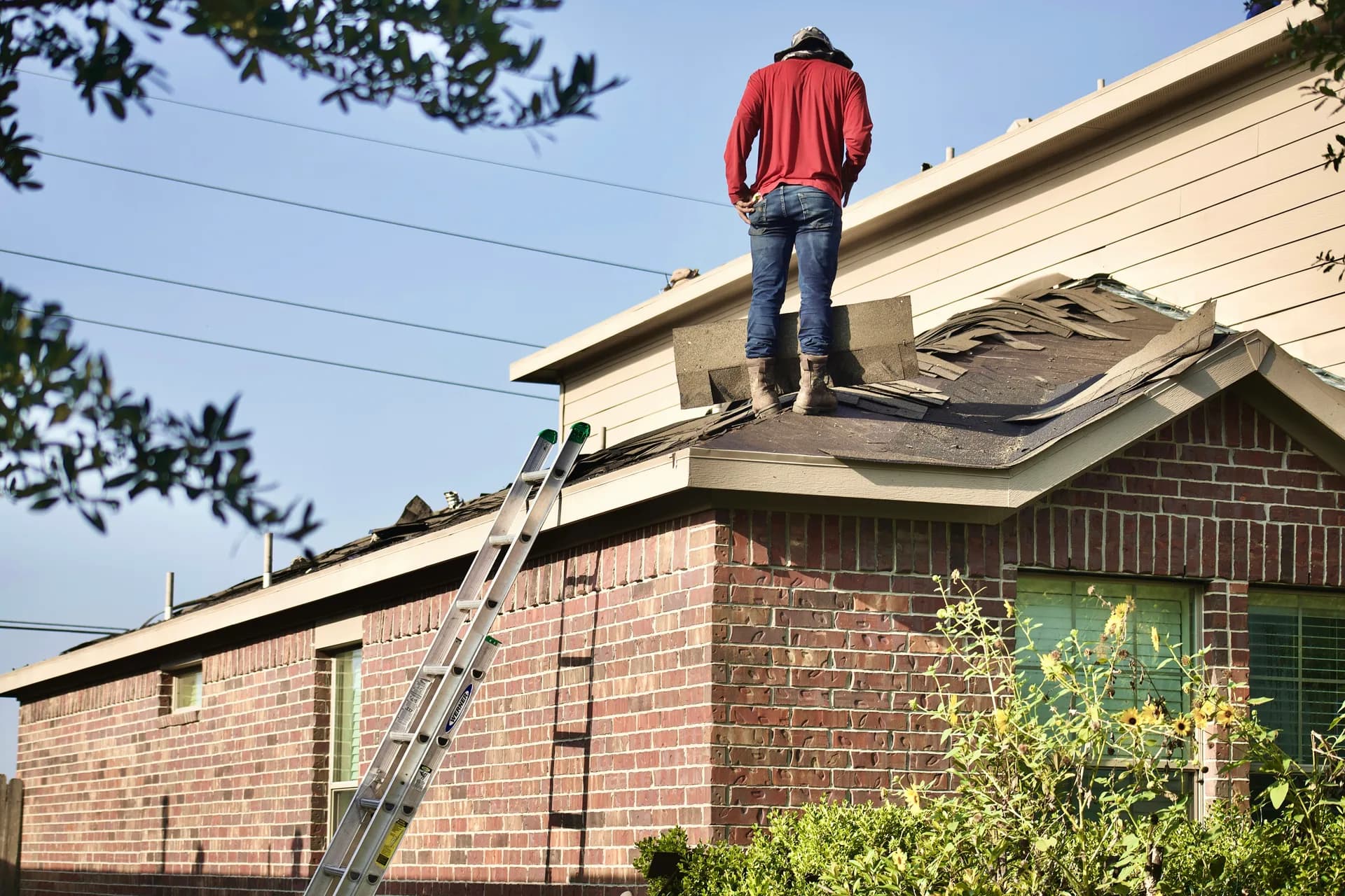Construction worker in red shirt standing on brick house roof doing roofing work with ladder visible