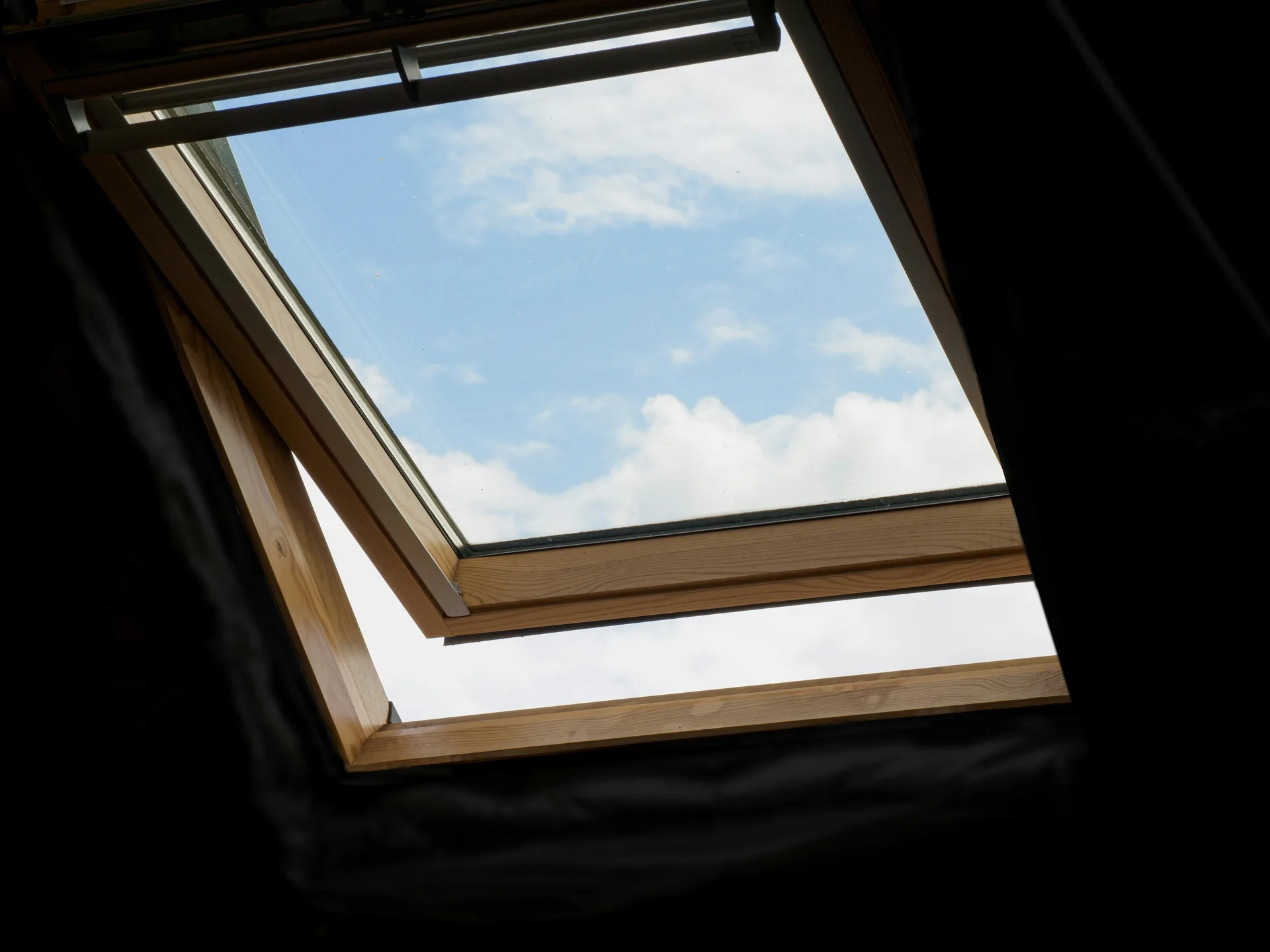 Open wooden-framed skylight window showing blue sky with white clouds from inside attic room