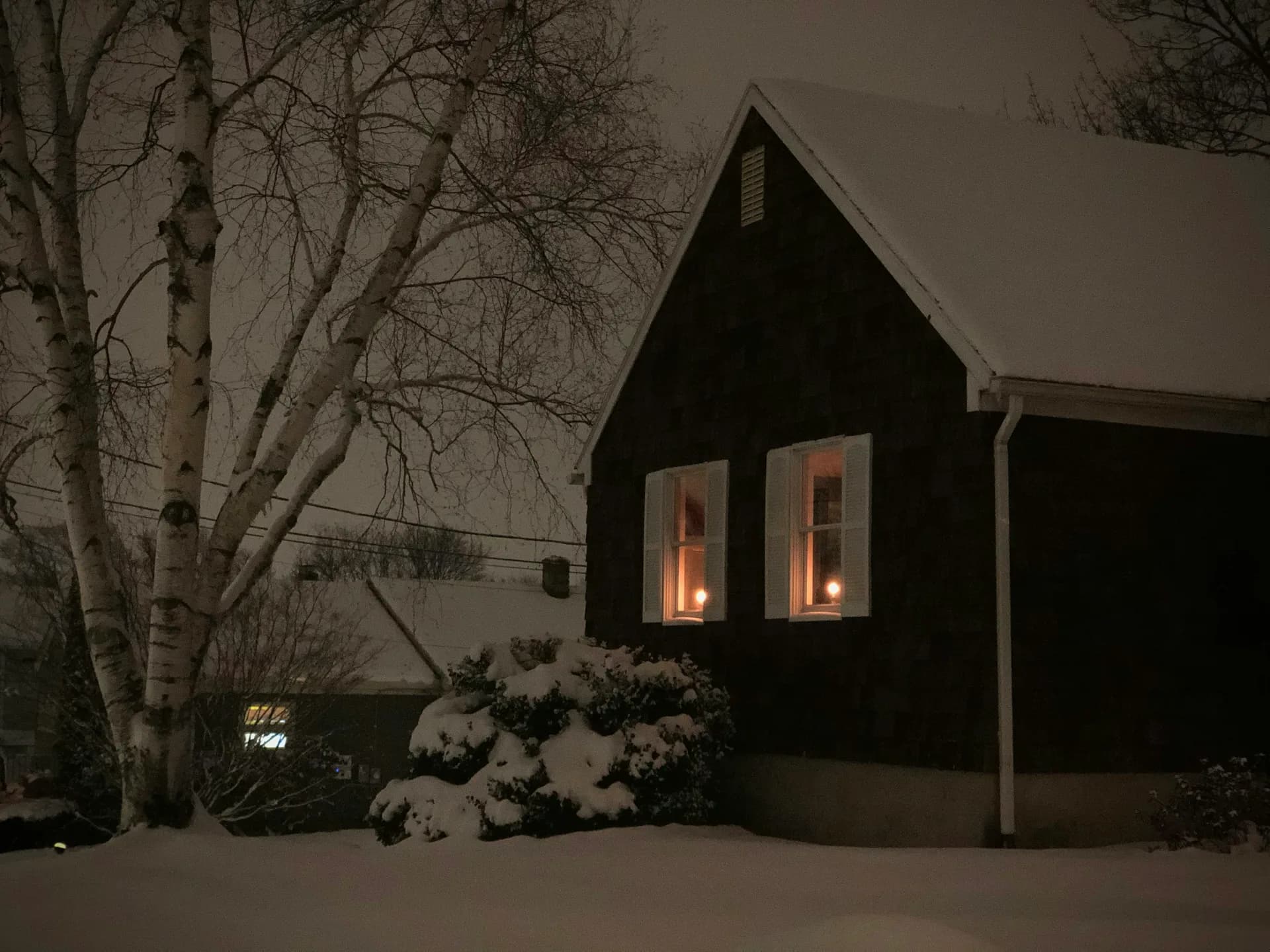 Snow-covered house with glowing windows on winter evening, surrounded by bare birch trees