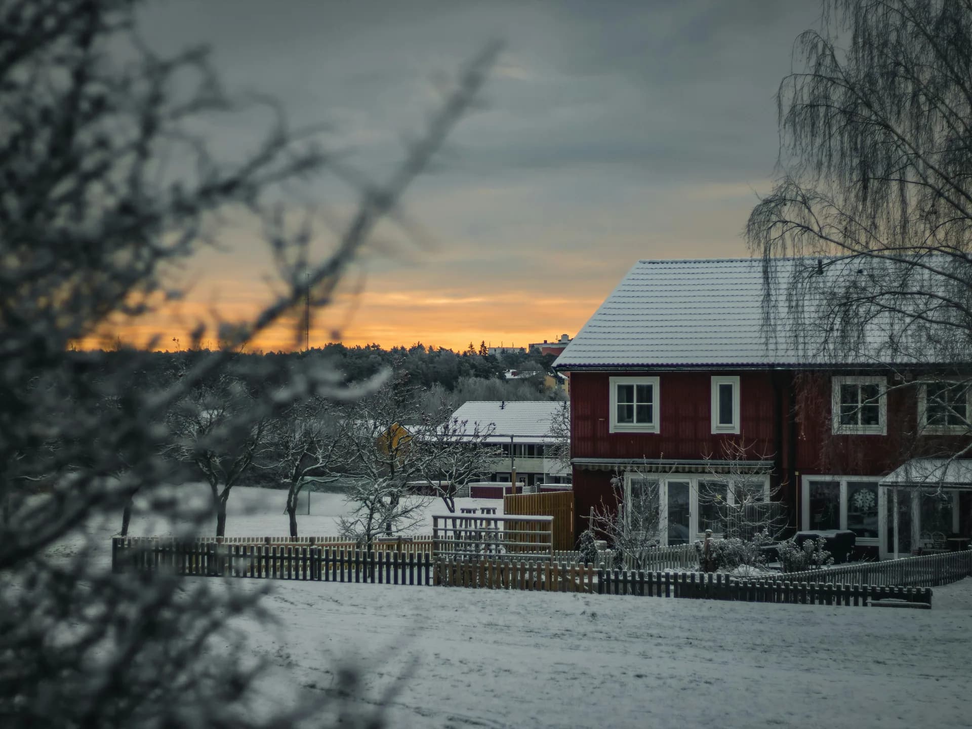 Red wooden house in snowy winter landscape at sunset with bare trees and wooden fence