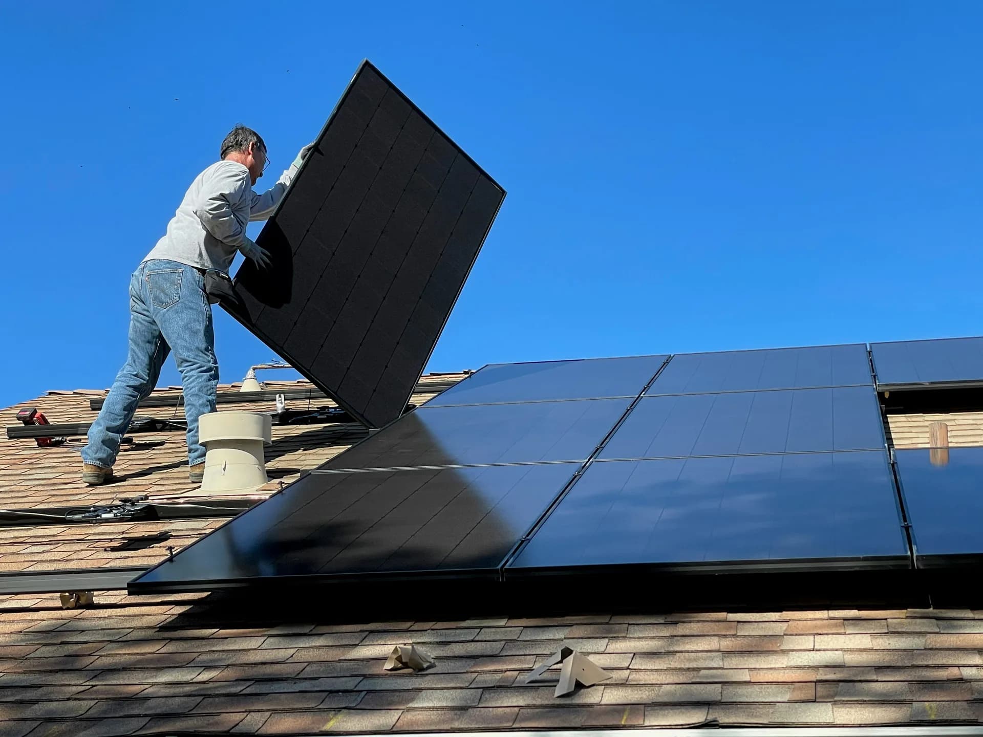 Worker installing black solar panels on residential roof with brown shingles under blue sky