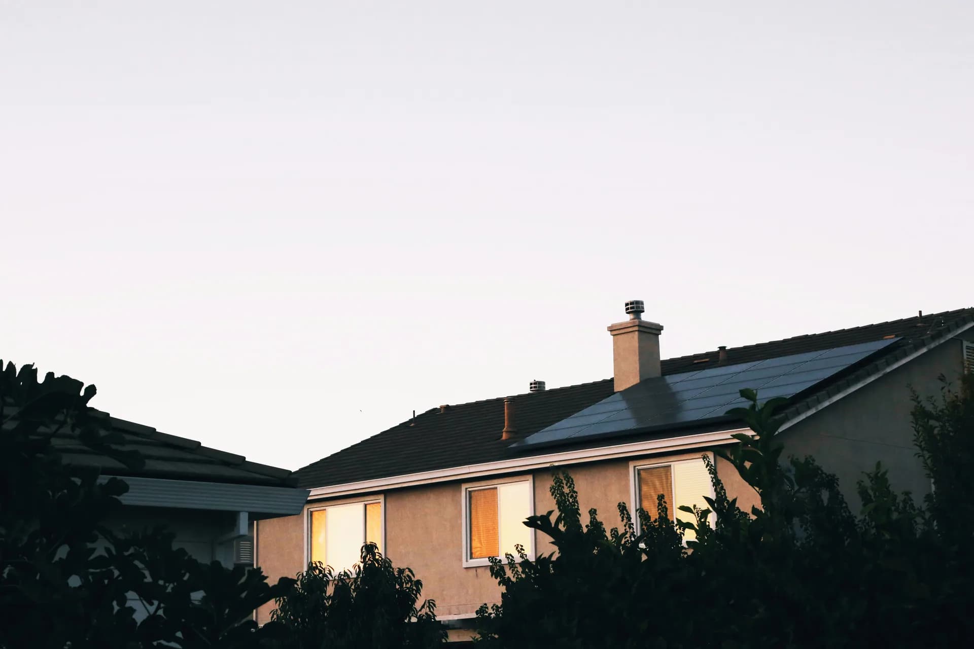 House at dusk with solar panels on roof and warm window lighting, surrounded by tropical plants