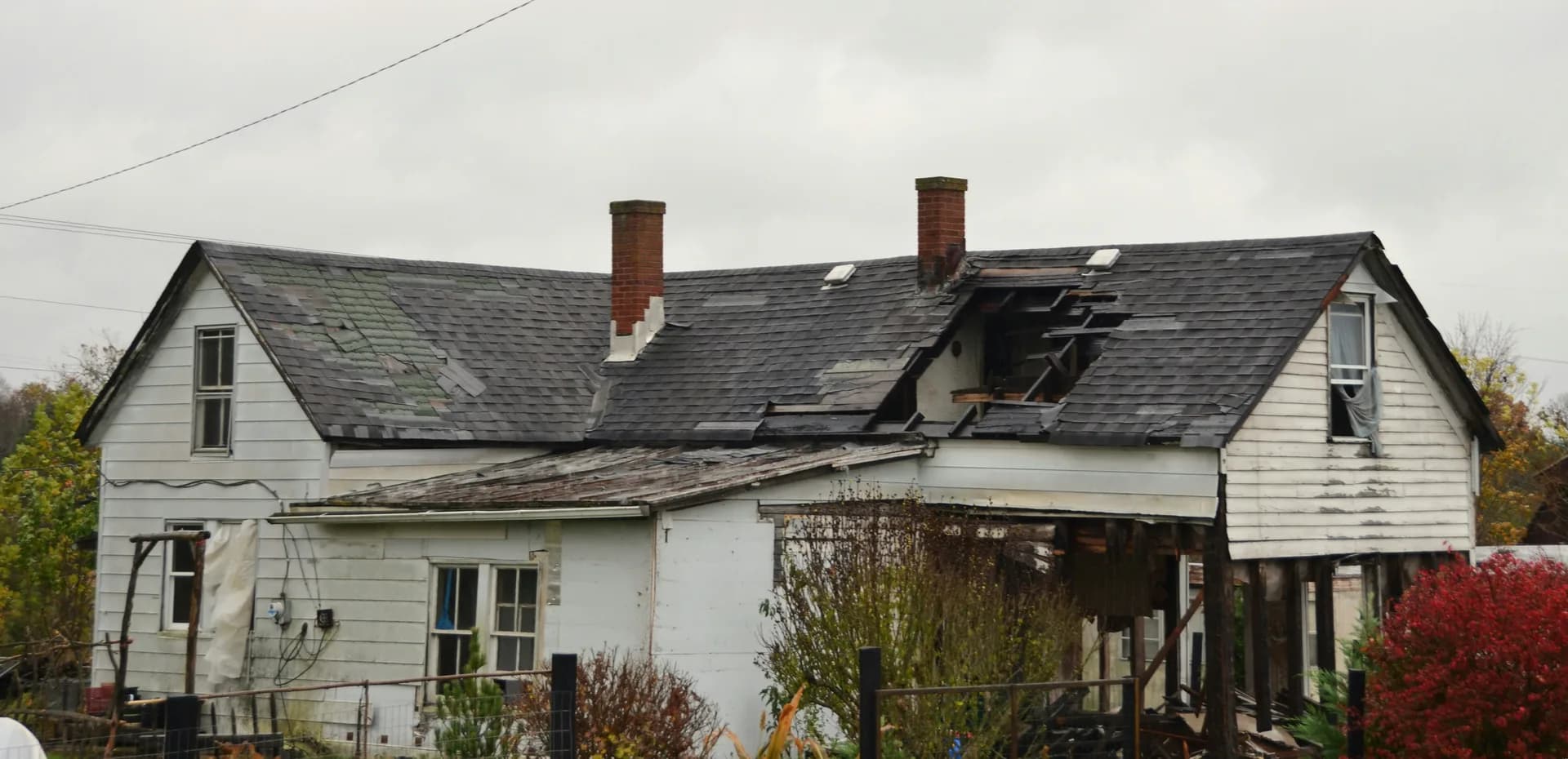 Fire-damaged white wooden house with collapsed roof sections, brick chimneys, and overgrown yard