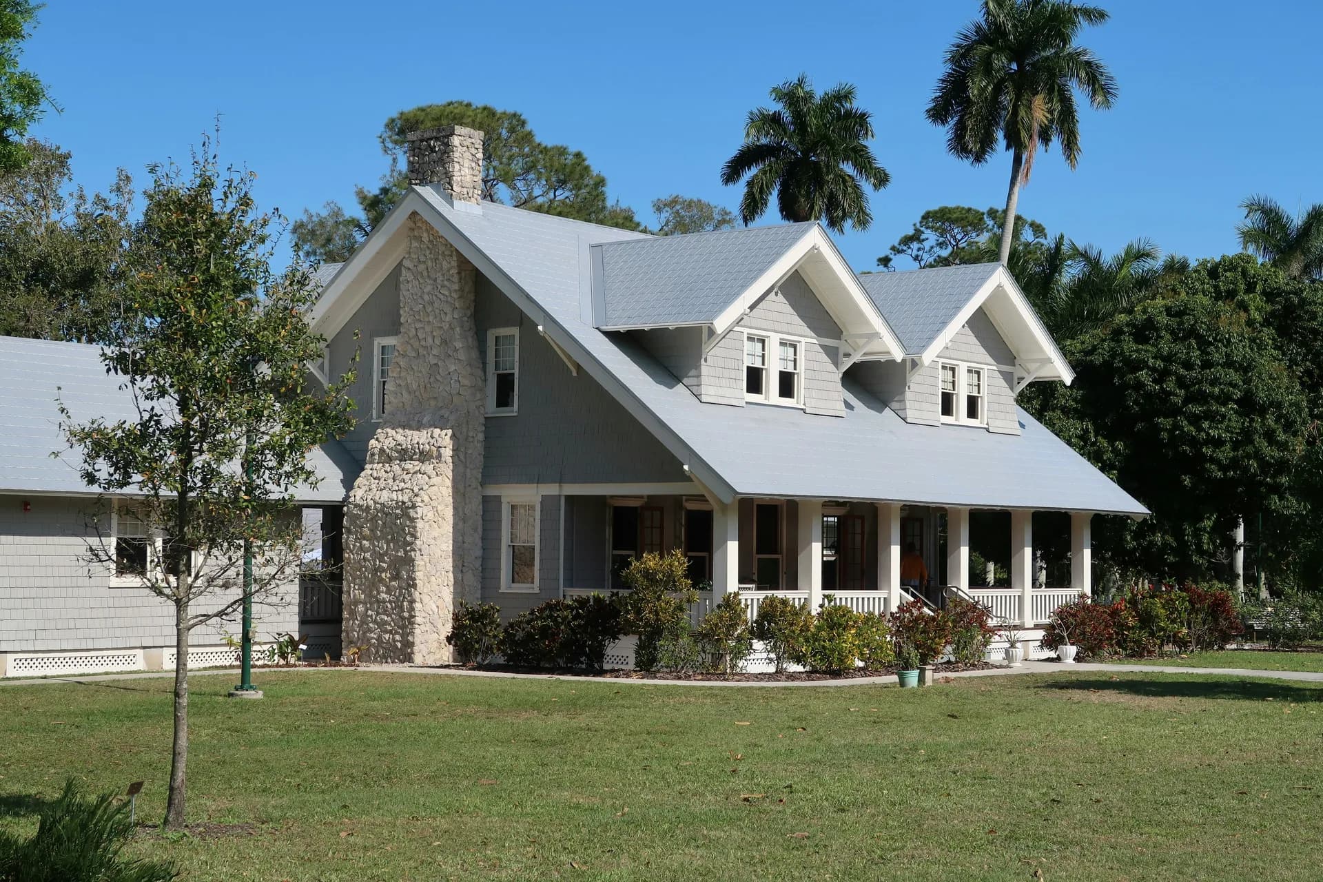 Gray shingled house with white wraparound porch, dormer windows, and tropical landscaping including palm trees
