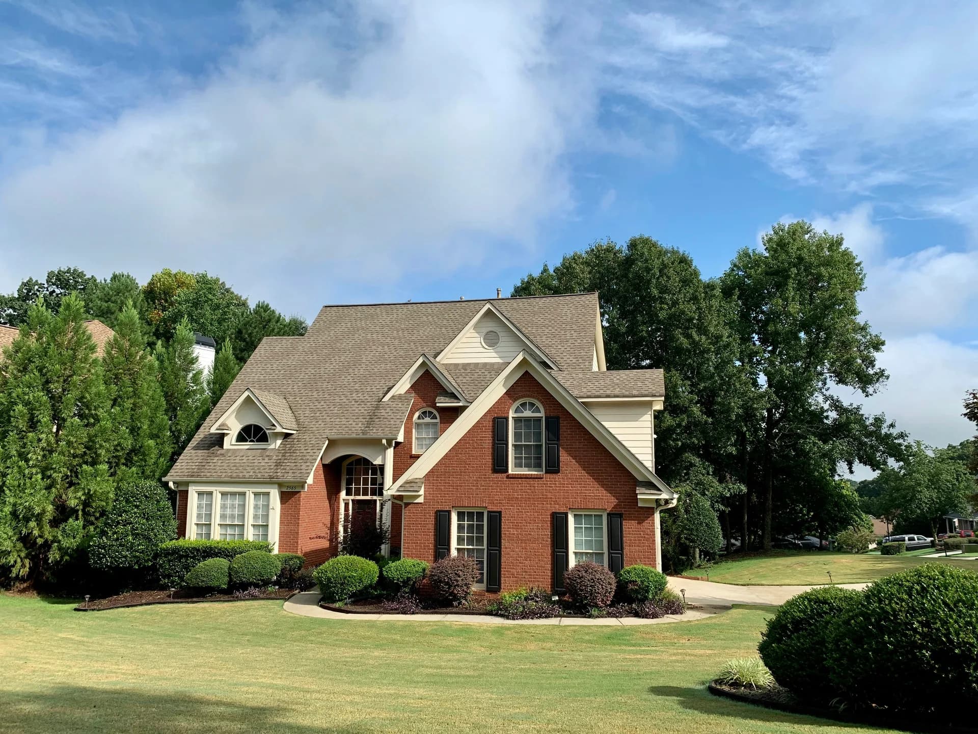 Two-story brick house with cream trim and dark shutters on manicured lawn with landscaping and mature trees