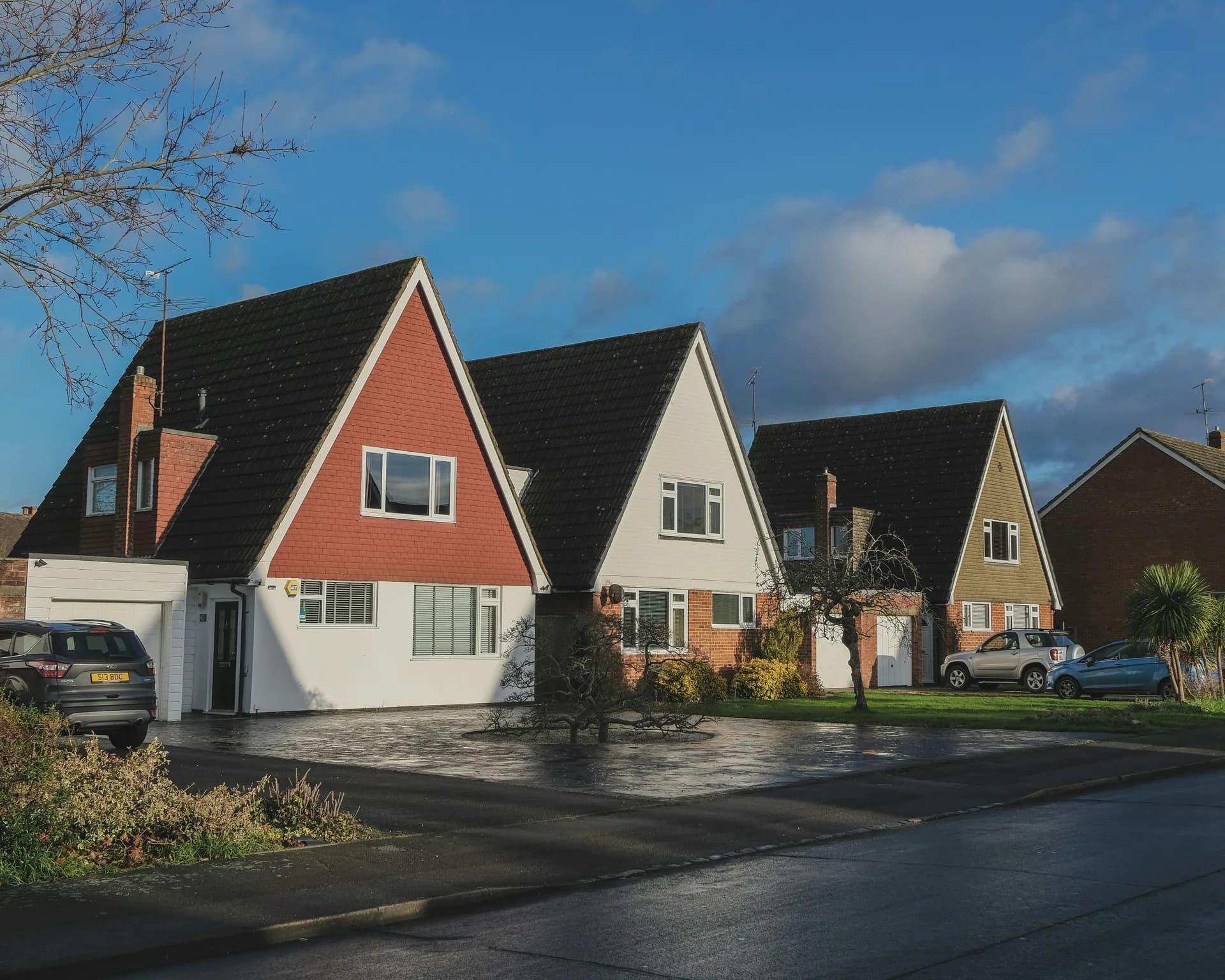 Row of English suburban houses with gabled roofs, mixed brick and painted exteriors, driveways and cars on residential street